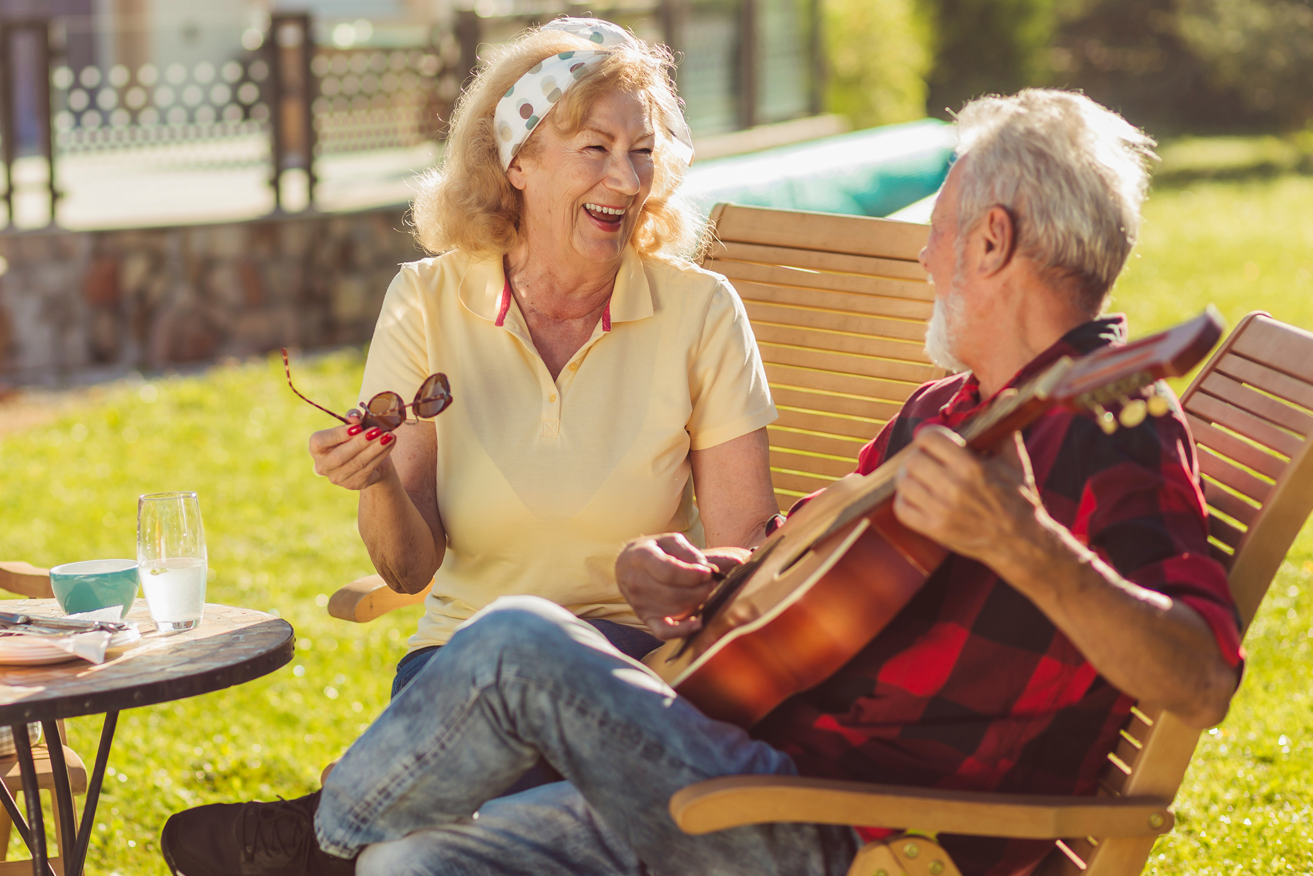 Cheerful senior couple having an outdoor lunch in the backyard, sitting at the table, drinking beer and lemonade, playing the guitar, singing and relaxing on a sunny summer day
